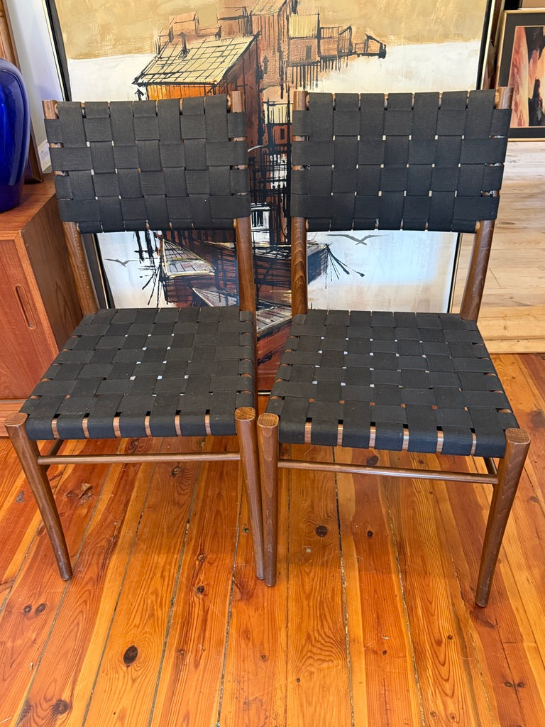 Two mid-century modern wooden dining chairs with black woven leather seats and backs, shown on a warm wood floor in front of framed artwork.